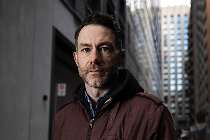 Brendan Kennedy, a man with short hair and a trimmed beard, stands in an urban alleyway, wearing a brown jacket over a hoodie.