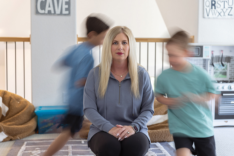 A woman with long blonde hair sits in a living room while two young children race around her in a blur.